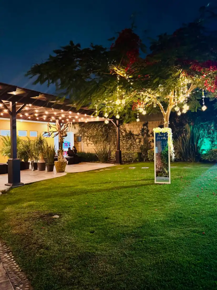 Jardín de Bonanza de noche, con luces cálidas colgando del árbol junto a la pérgola
