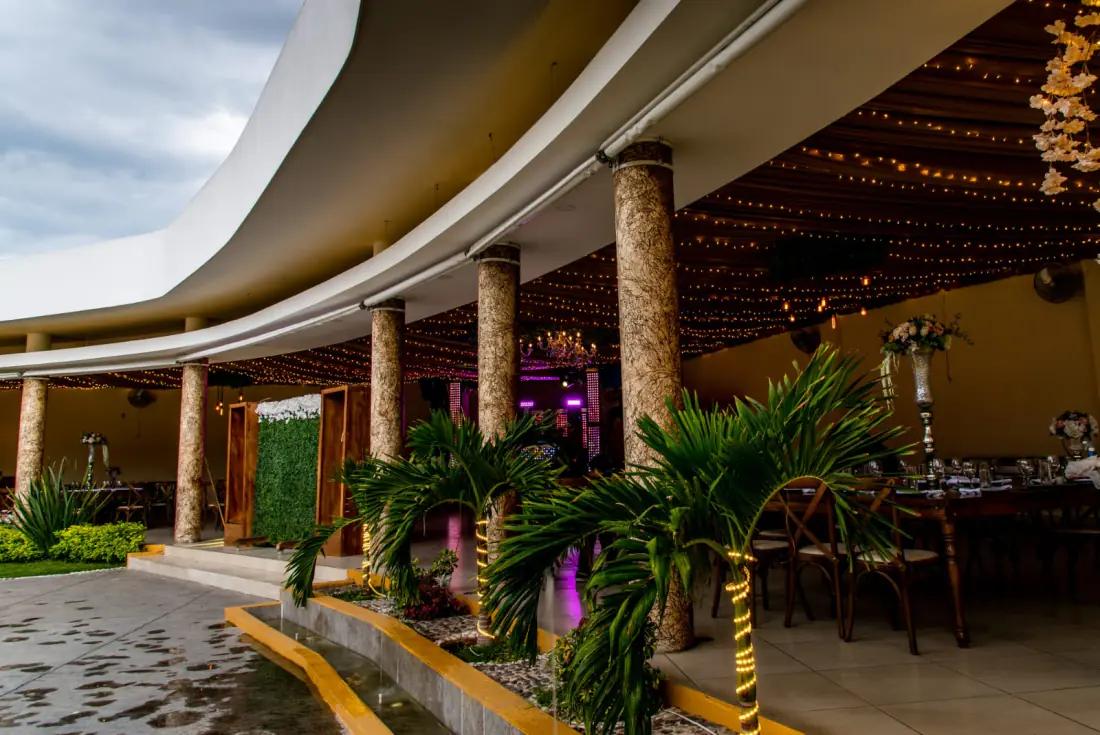 La terraza de Bonanza vista desde el jardín: marquesina curva, columnas de piedra y luces cálidas entre las palmeras